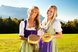 Zwei Personen in traditioneller Tracht präsentieren regionale Käsespezialitäten auf einer Wiese vor einer Bergkulisse