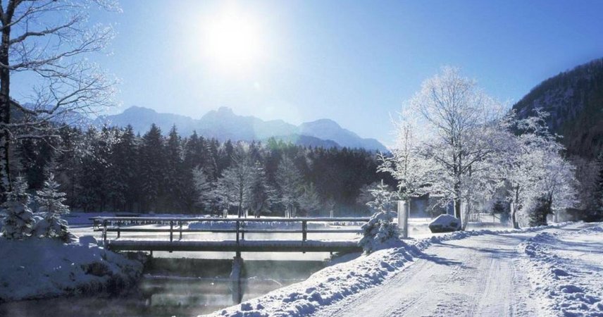 Verschneite Winterlandschaft mit Weg, Bach und Brücke vor einer Bergkulisse