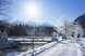 Winterliche Parklandschaft mit verschneitem Weg, Bach und Brücke vor einer beeindruckenden Bergkulisse