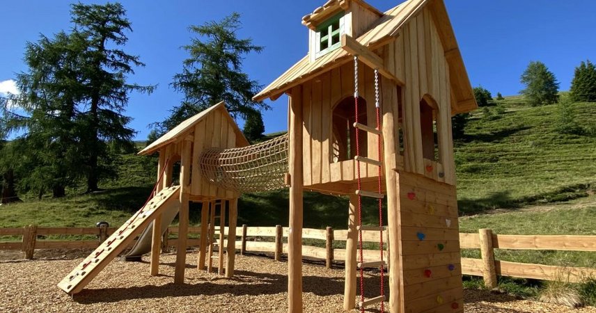Holzspielplatz mit mehreren Spieltürmen, Rutsche und Kletterelementen auf einer Wiese in sonniger Umgebung