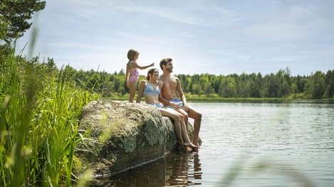 Drei Personen sitzen auf einem Felsen am Seeufer und halten die Füße ins Wasser