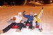Eine Gruppe von Menschen sitzt nachts auf bunten Schlitten im Schnee und jubelt auf einer beleuchteten Piste