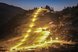 Eine nächtliche Skipiste, die von warmen Lichtern beleuchtet wird und sich durch eine dunkle Berglandschaft zieht